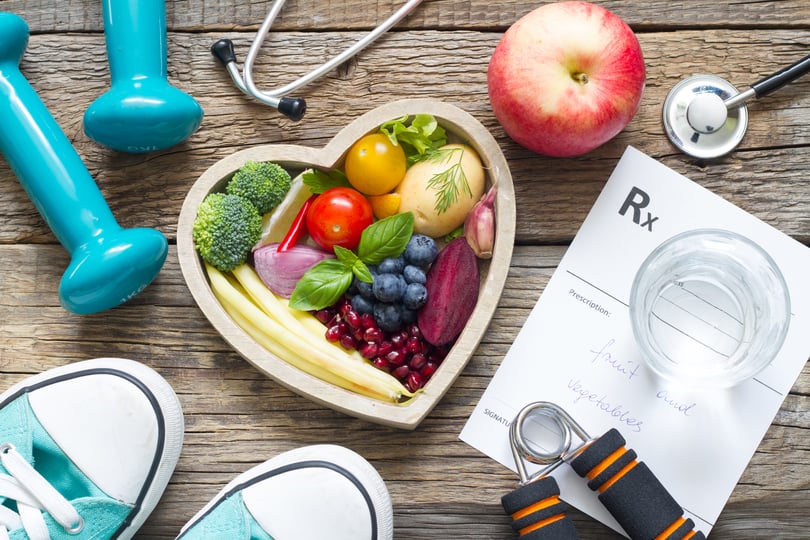 A wooden heart-shaped bowl full of fresh fruits and vegetables, sitting on a table next to exercise equipment, a glass of water, and a doctor’s note reading “fruit and vegetables.”