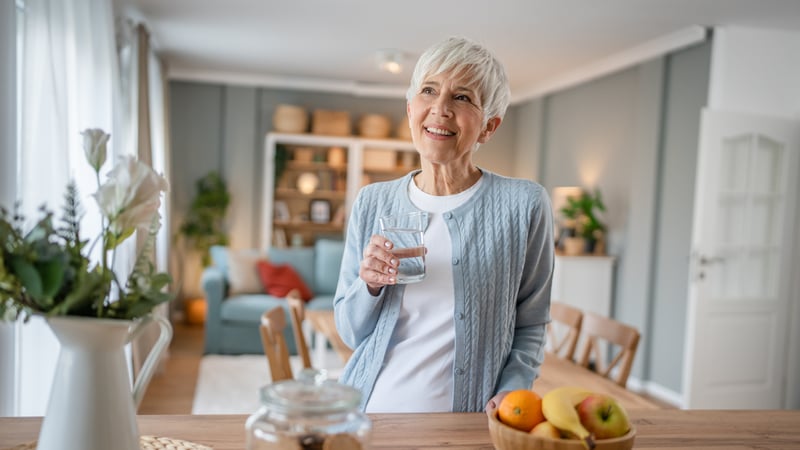 A senior woman smiling while holding a cup of water in her dining room.